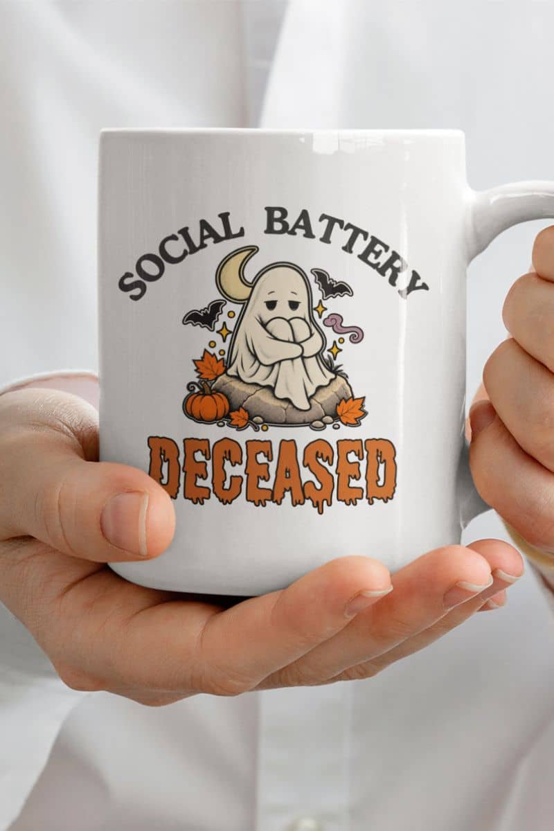 A person holding a ghost coffee mug featuring the text 'Social Battery: Deceased' around a ghost surrounded by pumpkins and bats.