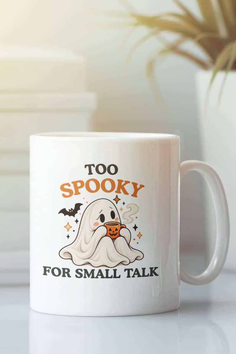 A white ghost coffee mug sits on a tabletop, featuring a shy ghost holding a pumpkin cup beneath the playful “Too Spooky for Small Talk” lettering.