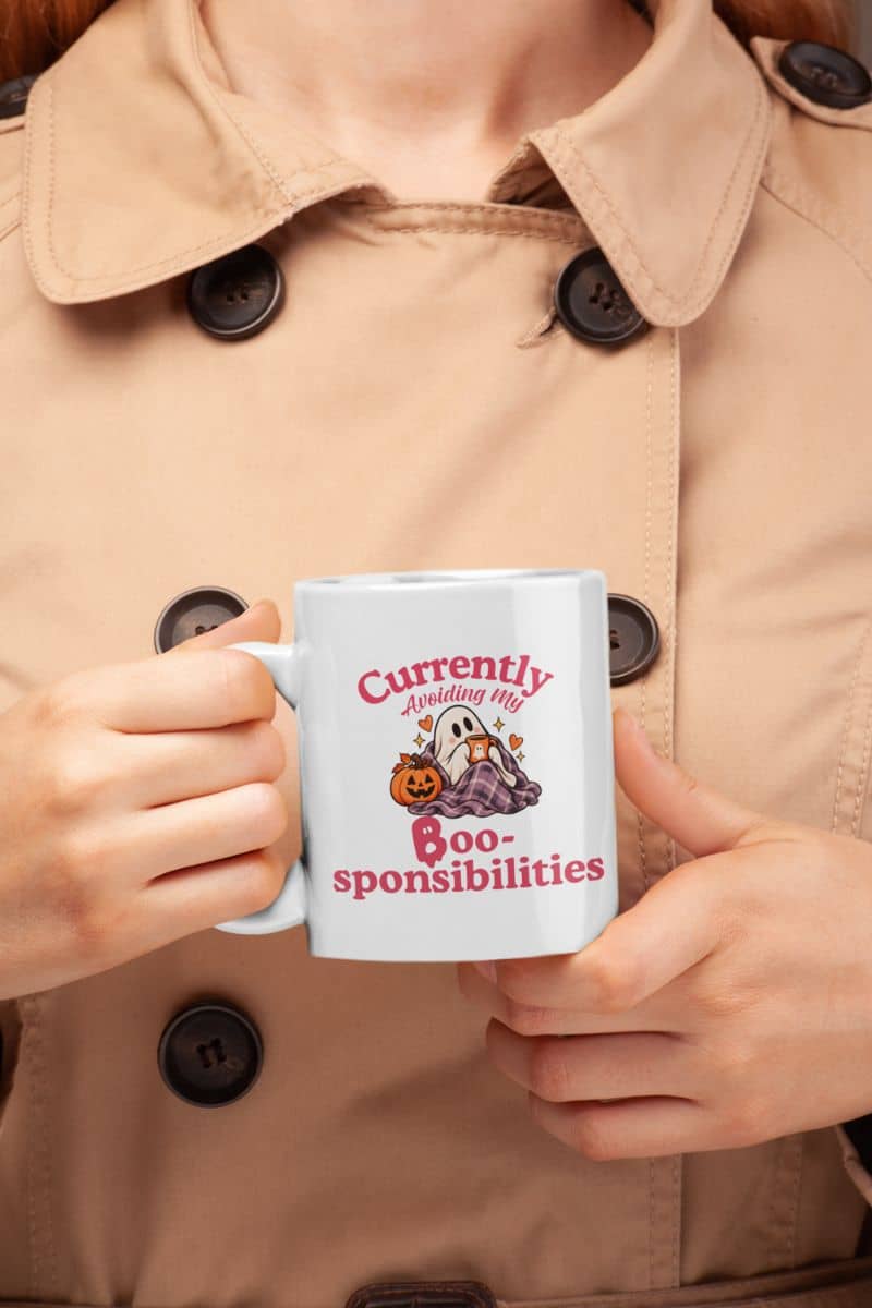 A person in a beige trench coat holding a ghost mug with a Halloween-themed graphic that reads 'Currently Avoiding My Boo-sponsibilities' alongside a ghost, pumpkin illustration.