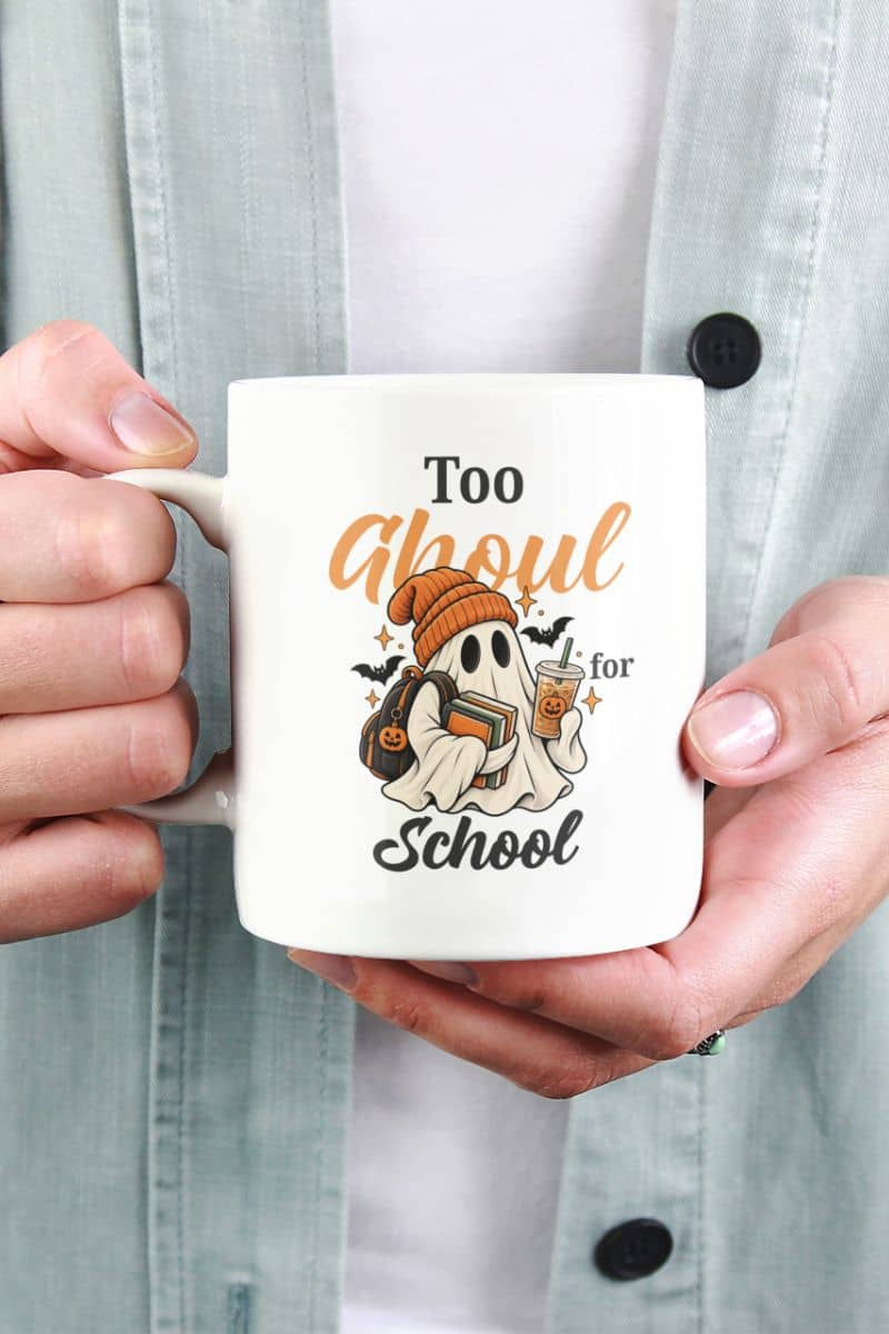 A person holding a white ghost mug with a Halloween-themed graphic that reads 'Too Ghoul for School' in orange and black text, featuring a ghost holding books and coffee