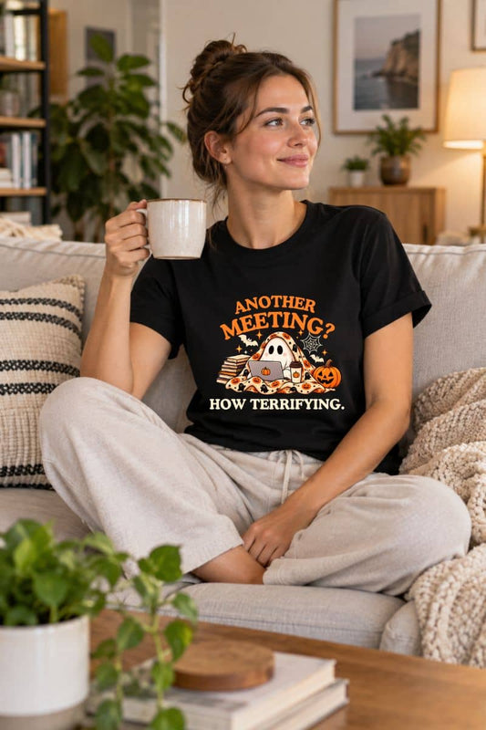 A woman sitting on a couch in a cozy living room, wearing a ghost shirt that reads 'Another Meeting? How Terrifying.' with a Halloween-themed illustration, and holding a white mug.