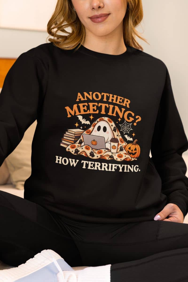 A woman wearing  a ghost sweatshirt that reads 'Another Meeting? How Terrifying.' with a ghost sitting on a sofa
