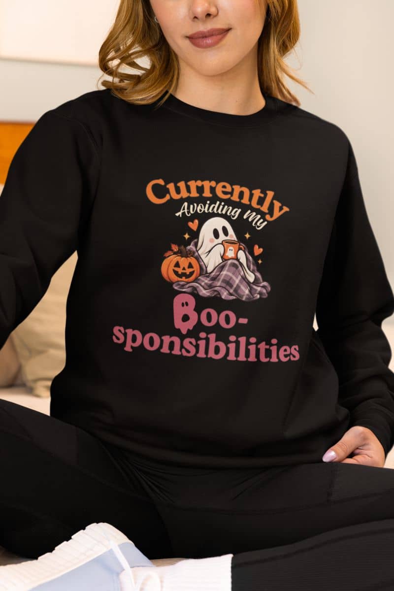 A woman wearing a ghost sweatshirt with a Halloween graphic that reads 'Currently Avoiding My Boo-sponsibilities' in orange and pink text, featuring a ghost, pumpkin, and plaid blanket.