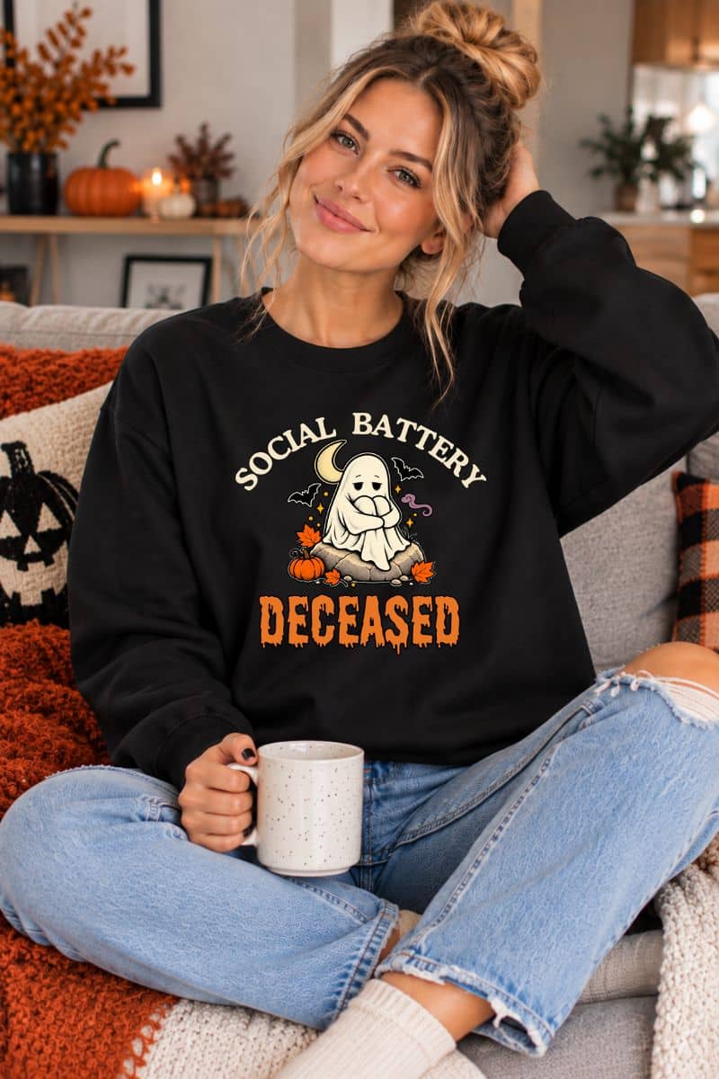 A woman wearing a ghost sweatshirt featuring the text 'Social Battery: Deceased' with a ghost sitting around decoritive items