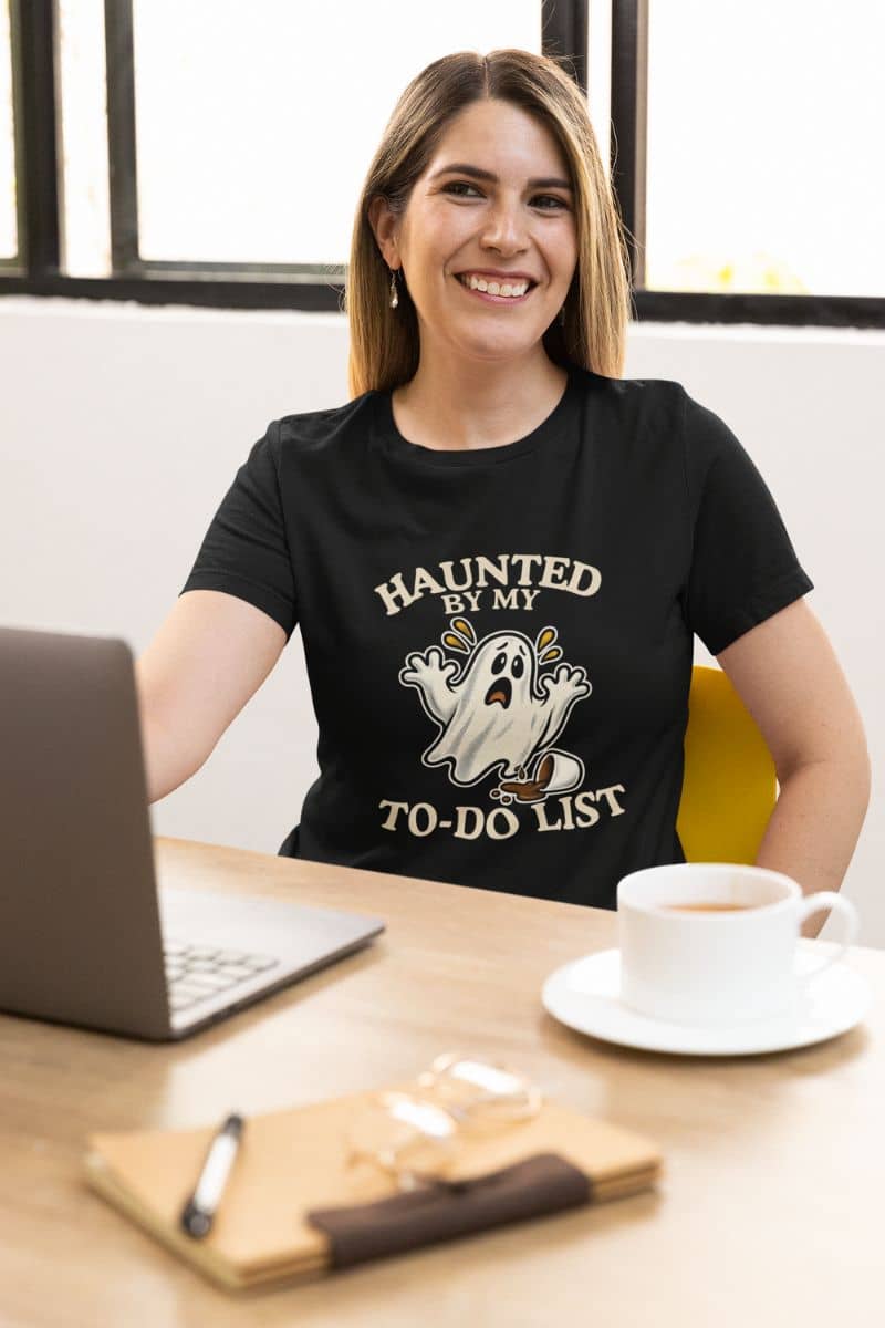 A woman wearing a ghost t shirt featuring a “Haunted by My To-Do List” funny ghost, sitting at the desk with a laptop, a note book and a cup of tea