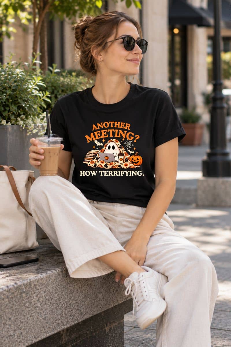 A woman sitting outdoors on a stone bench wearing a ghost t shirt with a Halloween-themed design and the text 'Another Meeting? How Terrifying.' She is also wearing white pants, white sneakers, sunglasses, and holding an iced coffee.