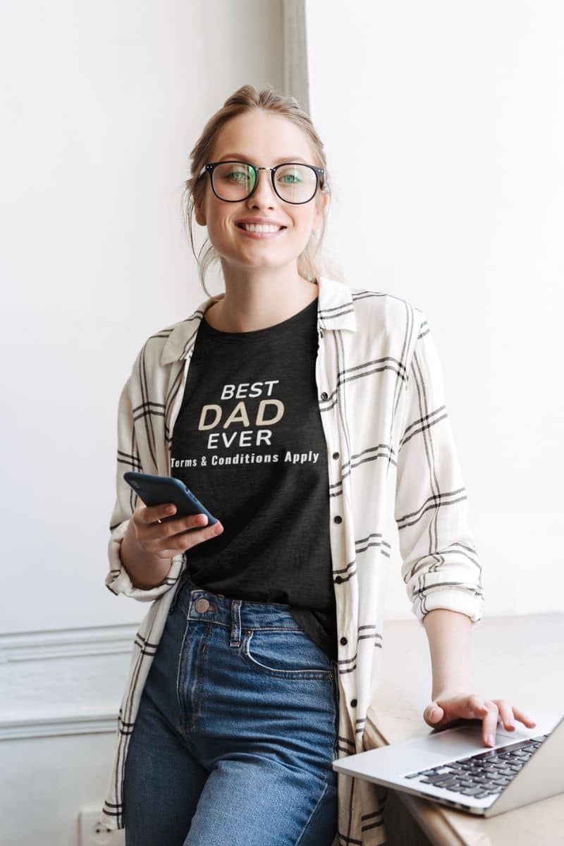gift for dad shirt with a woman wearing a black t-shirt with 'Best Dad Ever, Terms & Conditions Apply' text, holding a phone and using a laptop.