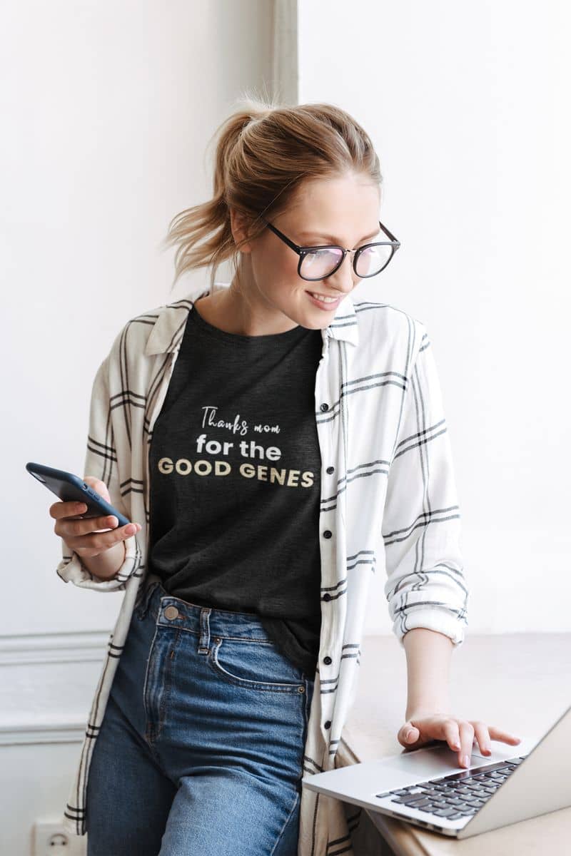 gift for mom shirt with a woman wearing a black t-shirt with text 'Thanks Mom for the Good Genes', using a laptop and smartphone indoors.