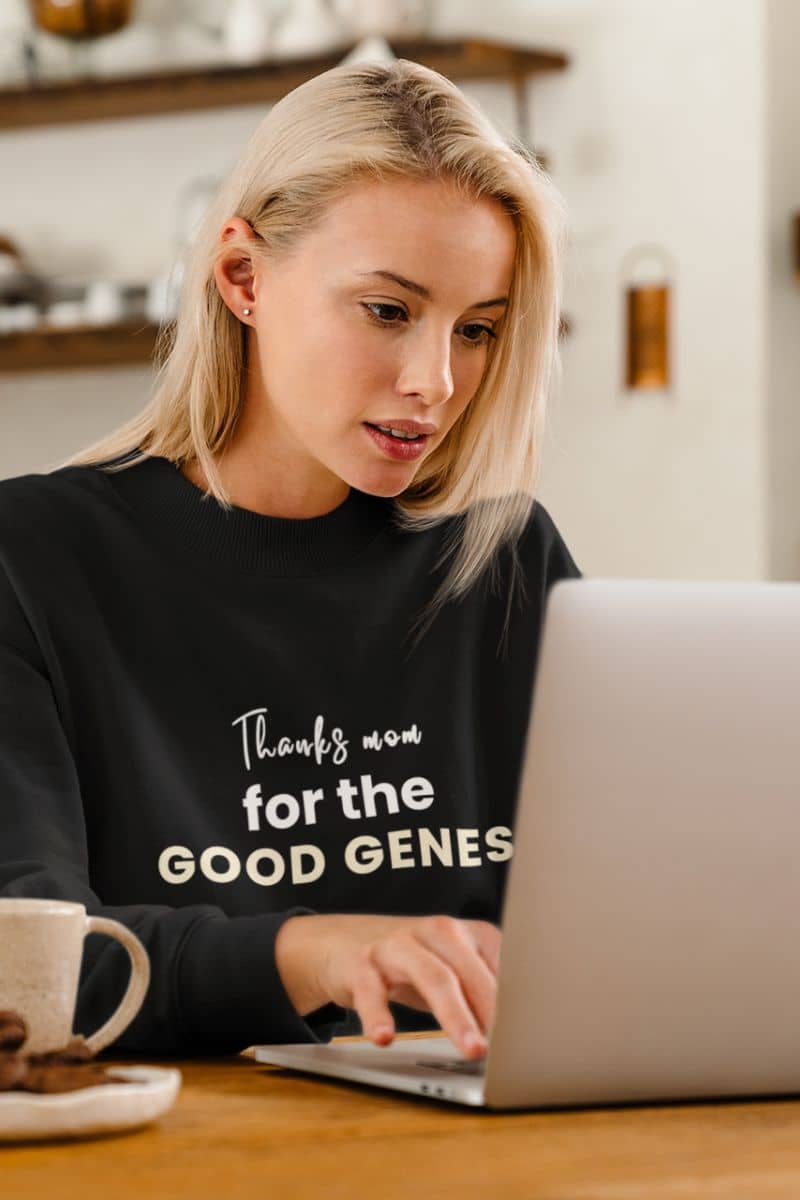 gift for mom sweatshirt with a woman wearing a black sweatshirt with text 'Thanks Mom for the Good Genes', using a laptop at a table.
