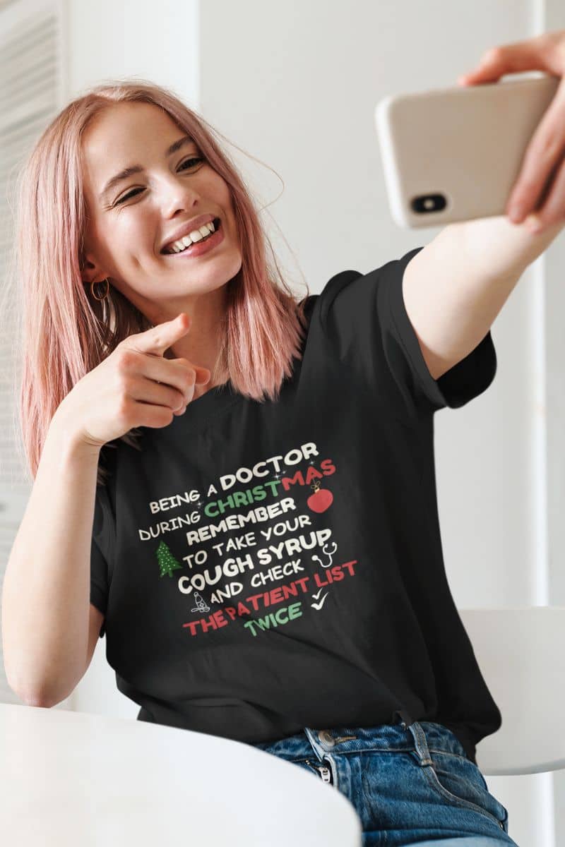 healthcare christmas shirt with a person wearing a black t-shirt with text 'Being a doctor during Christmas, remember to take your cough syrup and check the patient list twice', taking a selfie.