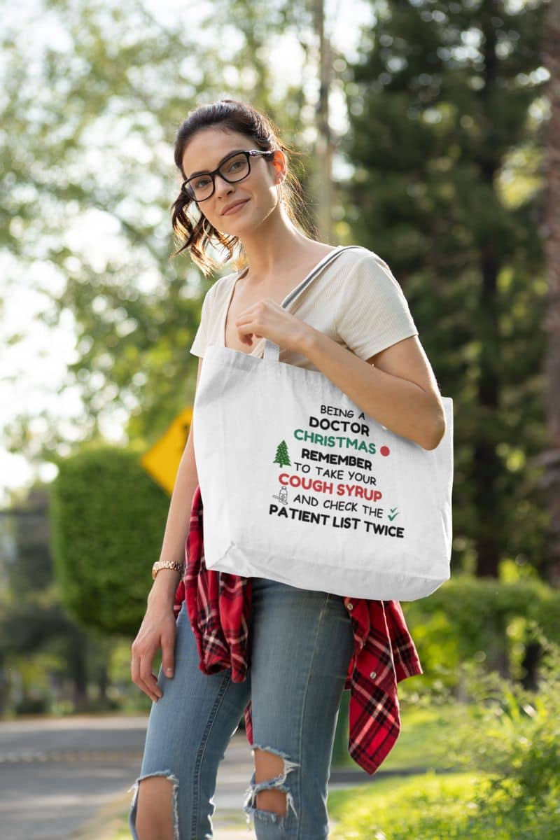 healthcare christmas shopper with a woman holding a tote bag with text 'Being a doctor during Christmas, remember to take your cough syrup and check the patient list twice' outdoors.