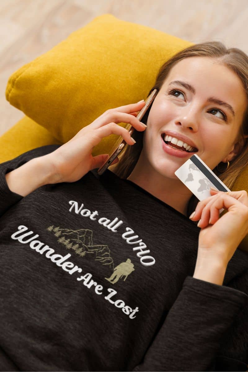hiking lifestyle sweatshirt with a woman wearing a black sweatshirt with text 'Not all who wander are lost', holding a phone and card, lying on a yellow pillow.