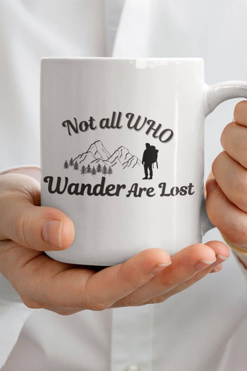 hiking mug gift with a white mug with 'Not all Who Wander Are Lost' text and mountain design held by hands against a white background
