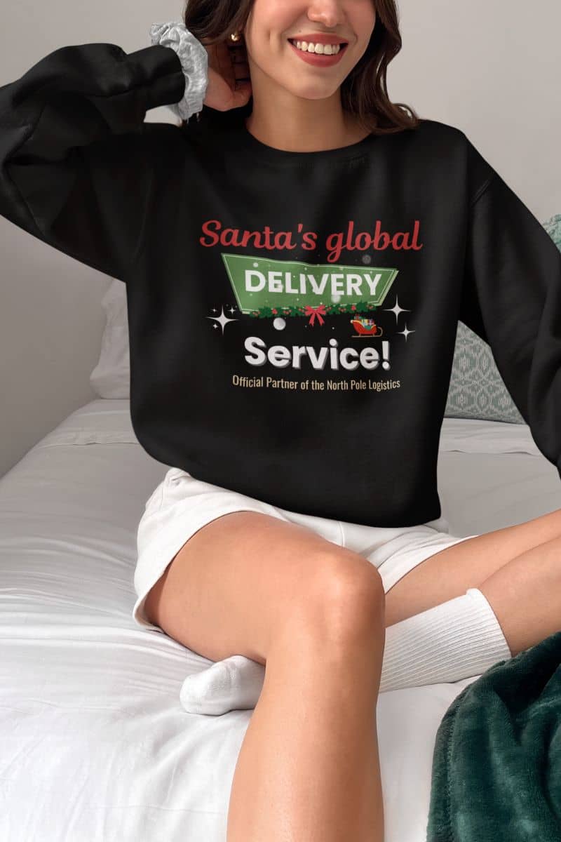 holiday crewneck with a person wearing a black sweatshirt with Christmas-themed text sitting on a bed.