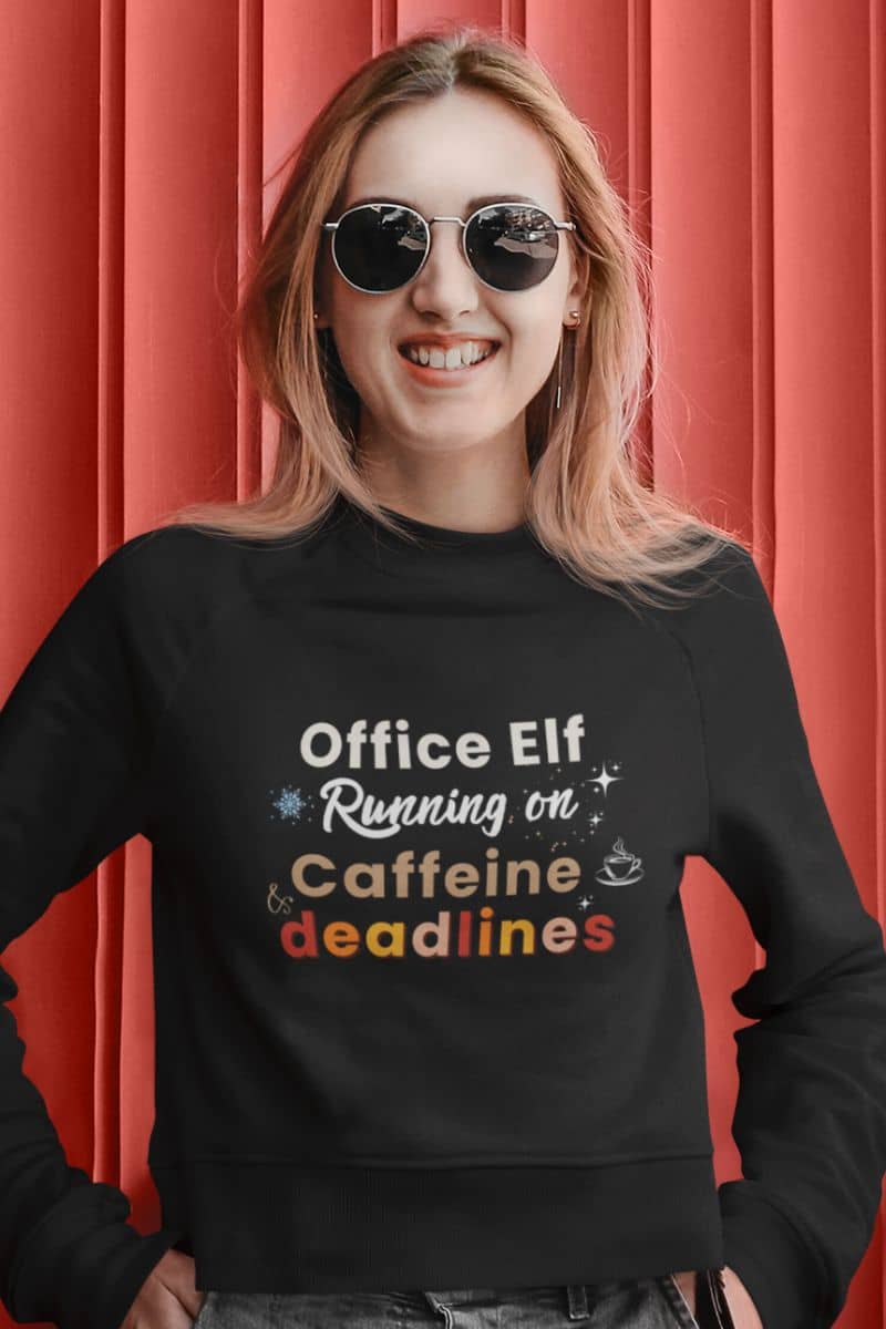 holiday office sweater with a person wearing a black sweatshirt with humorous text 'Office Elf, Running on Caffeine & Deadlines' against a red background