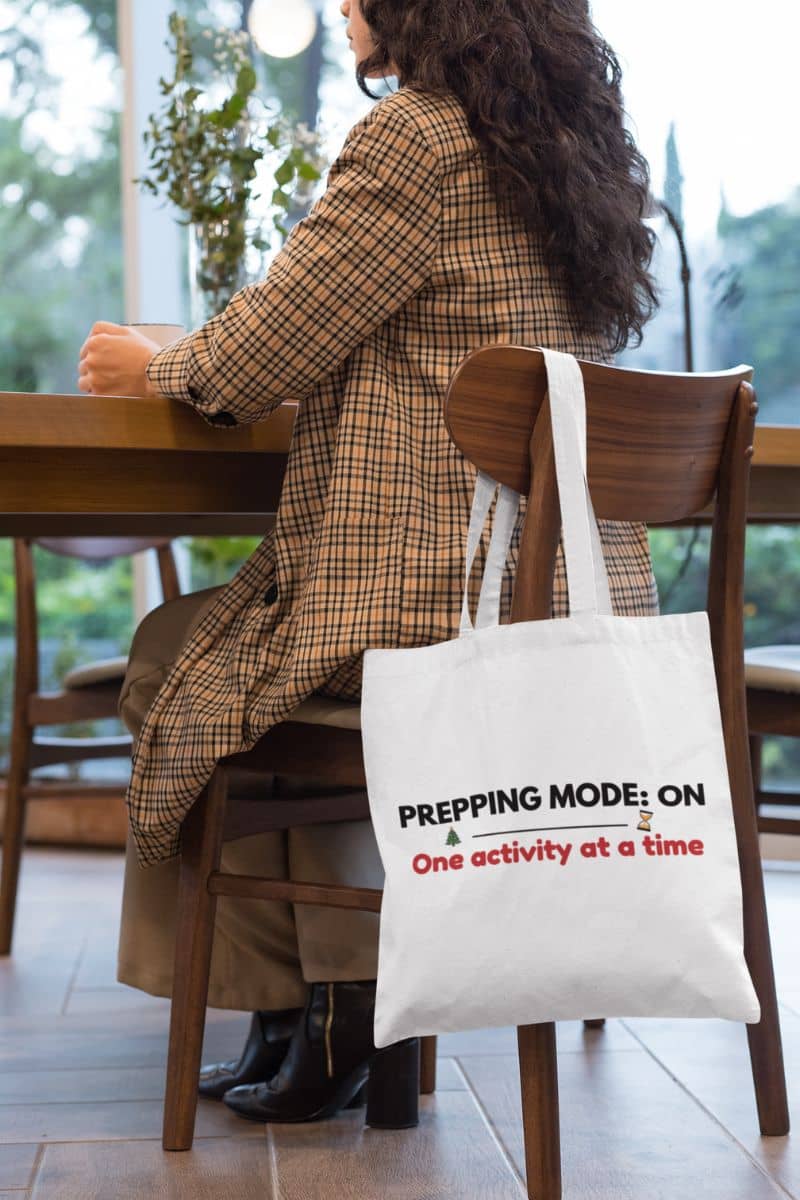 holiday planning tote bag with a person sitting at a table with a tote bag featuring text 'Prepping Mode: ON – One activity at a time', in an indoor setting.