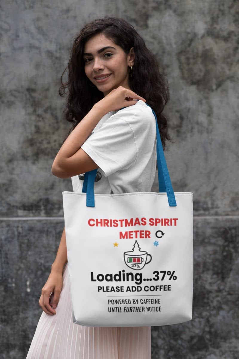 holiday shopping bag with a woman holding a tote bag with text 'Christmas Spirit Meter, Loading…37%. Please add coffee. Powered by caffeine until further notice' against a gray wall.