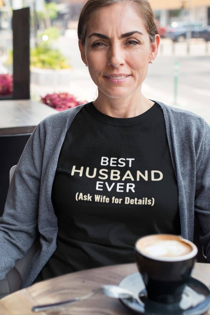 husband humor tee with a woman wearing a black t-shirt with text 'Best Husband Ever (Ask Wife for Details)', sitting at a table with a coffee cup.