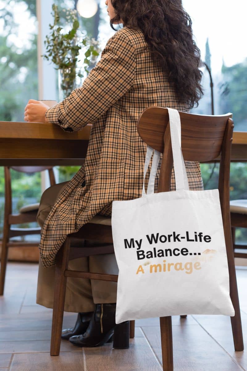 hustle joke tote with a person sitting at a table with a tote bag that says 'My Work-Life Balance... A Mirage' in a blurred indoor setting.