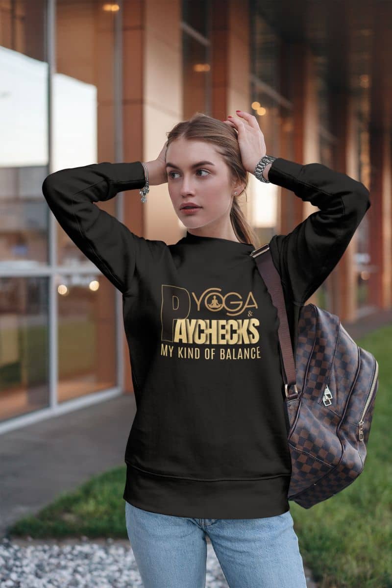 inspirational crewneck with a woman wearing a black hoodie with gold text 'Yoga & Paychecks – My Kind of Balance', standing outdoors.