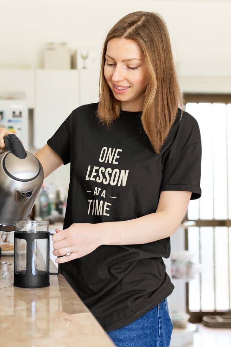 inspirational teacher shirt with a woman in a kitchen wearing a black t-shirt that reads 'ONE LESSON AT A TIME', pouring water from a kettle into a French press.