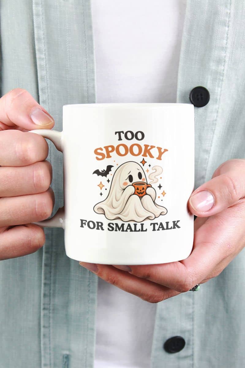 A person in a light blue shirt holds an introvert coffee mug featuring a shy ghost sipping from a pumpkin cup and the quote “Too Spooky for Small Talk.”