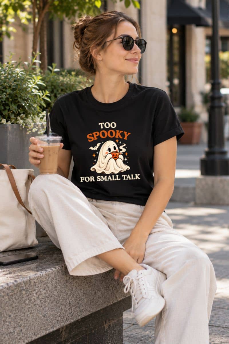 Woman wearing a black introvert Halloween shirt featuring a shy ghost holding coffee, sitting outdoors with an iced drink.