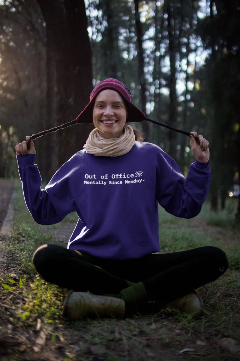 introvert mode hoodie with a person sitting in a forest wearing a purple sweatshirt with text 'Out of Office. Mentally Since Monday', holding a hat.