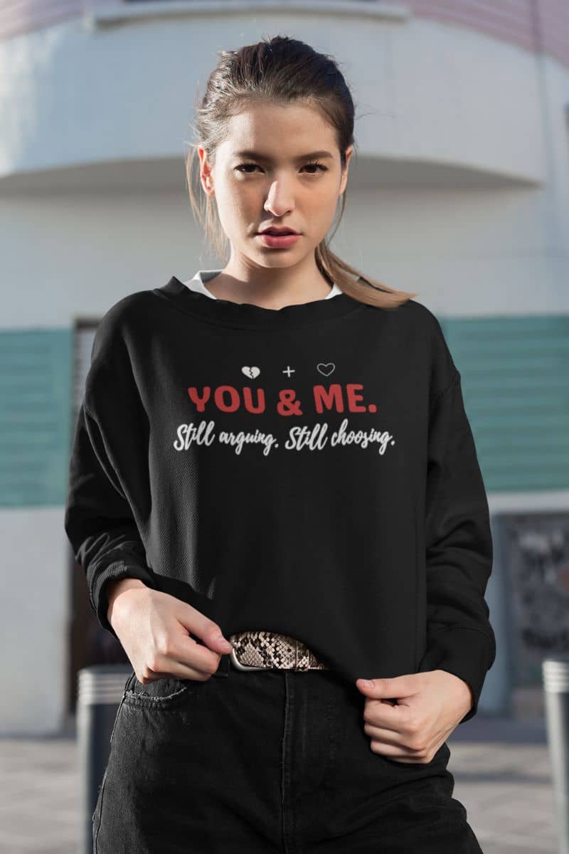 long lasting love sweatshirt with a woman wearing a black sweatshirt with text 'YOU & ME. Still arguing. Still choosing.', standing outdoors.