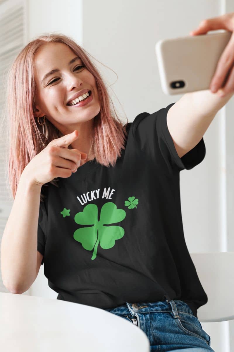 lucky charm t-shirt with a person wearing a black t-shirt with a green four-leaf clover design and 'Lucky Me' text, taking a selfie.