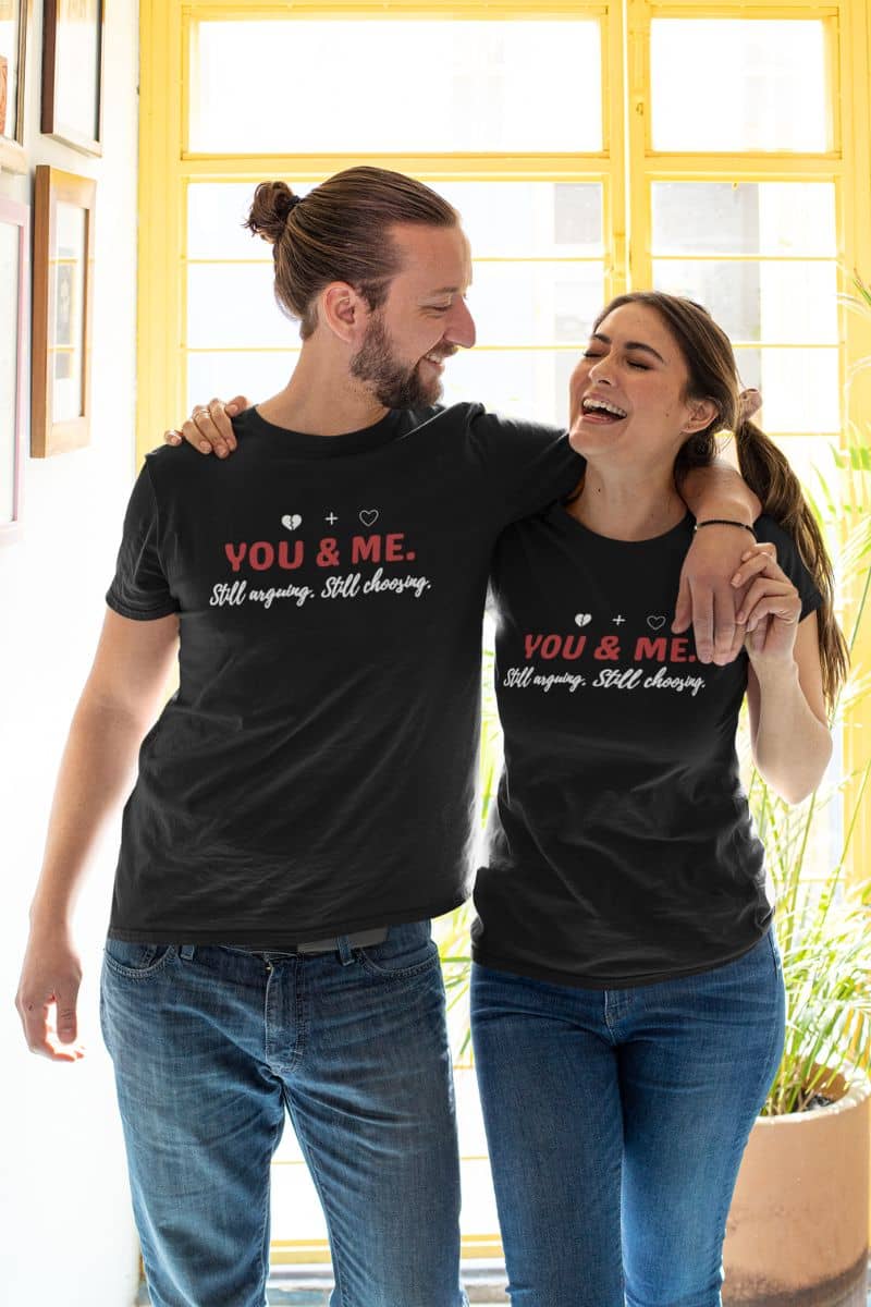 marriage humor t-shirt with two people wearing matching black t-shirts with 'YOU & ME. Still arguing. Still choosing.' text, standing together indoors.