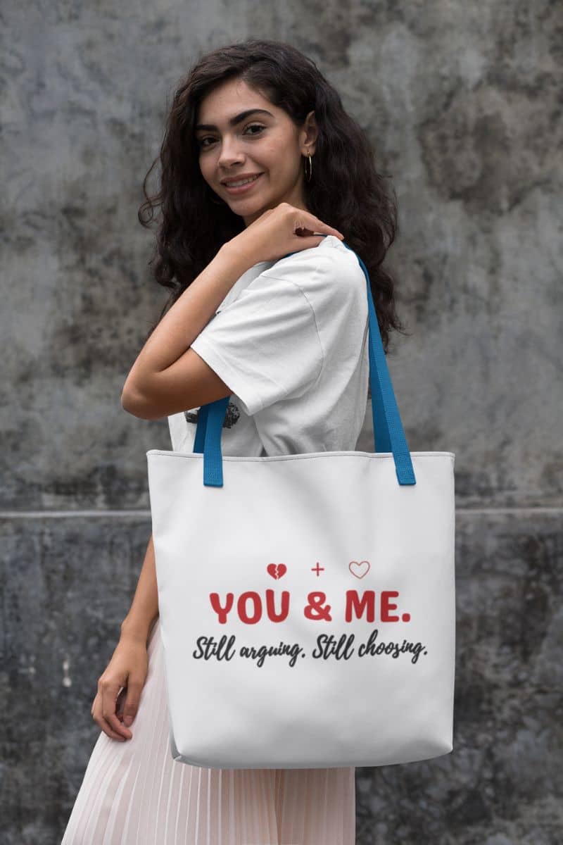 marriage humor tote with a woman holding a tote bag with 'You & Me. Still arguing. Still choosing.' text against a gray wall.