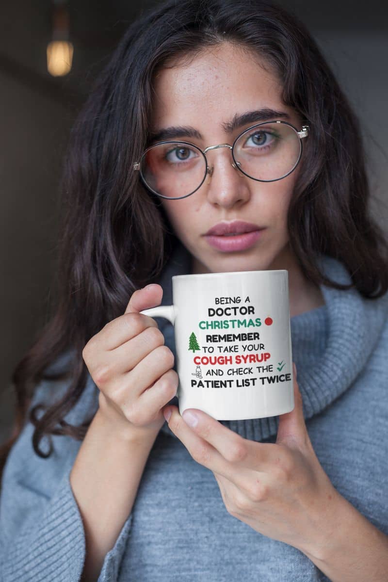 medical humor mug with a person holding a mug with text 'Being a doctor during Christmas, remember to take your cough syrup and check the patient list twice', wearing glasses and a gray sweater.