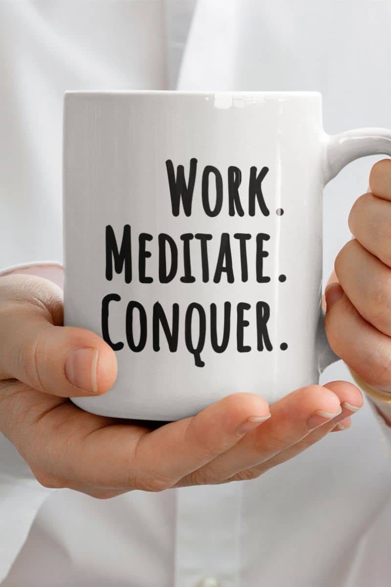 meditation desk mug with a white mug with 'Work. Meditate. Conquer.' text held by hands against a light background