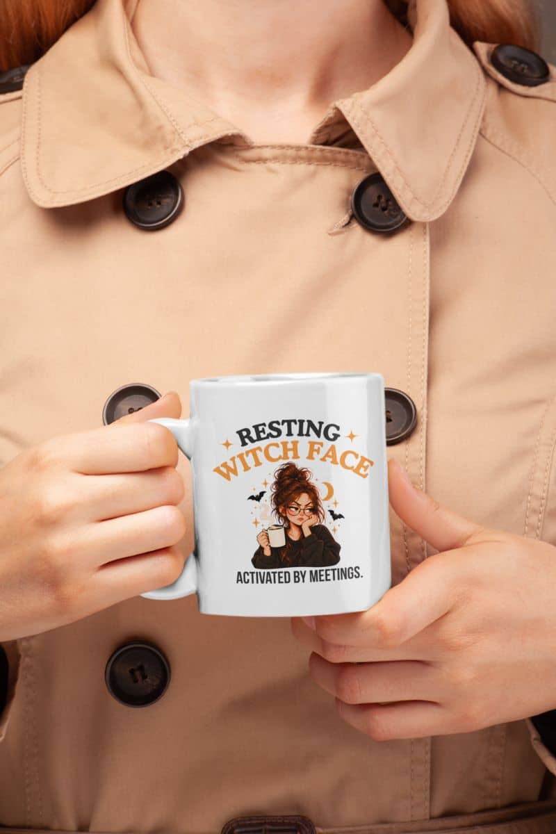 A person wearing a beige trench coat holding a meeting coffee mug with a Halloween graphic that reads 'Resting Witch Face. Activated by Meetings'.