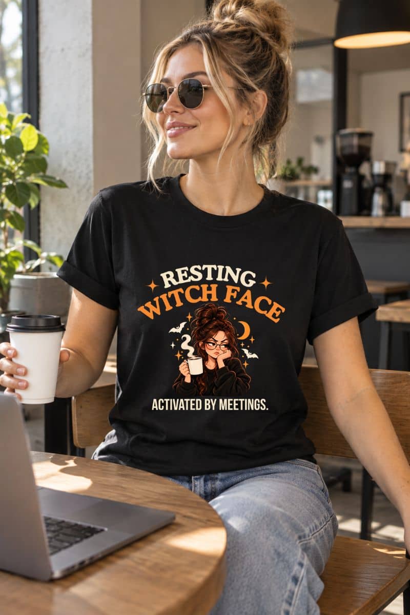 A woman wearing a meeting humor shirt with the text 'Resting Witch Face. Activated by Meetings' in orange and white, sitting in a cafe setting.