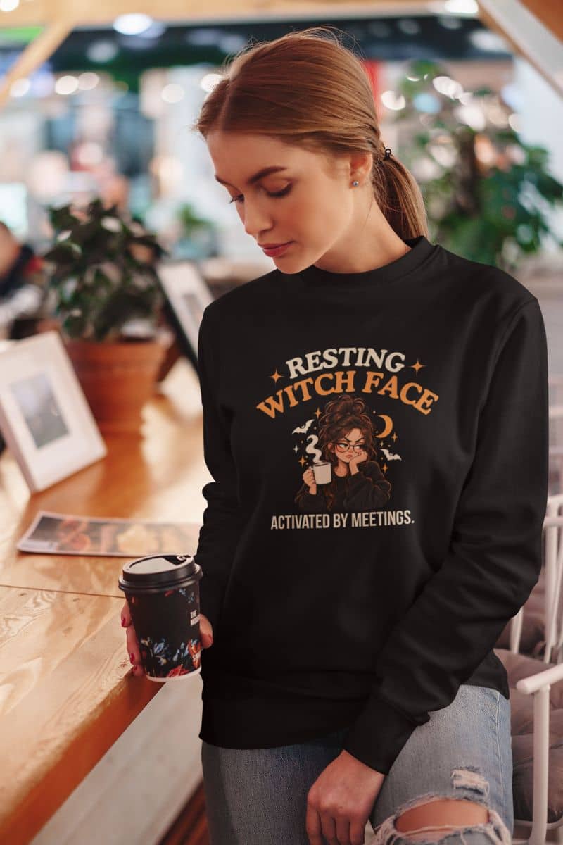 A woman standing at the coffee shop, wearing a sweatshirt featuring a "Resting Witch Face. Activated by Meetings." graphic with a woman's face 