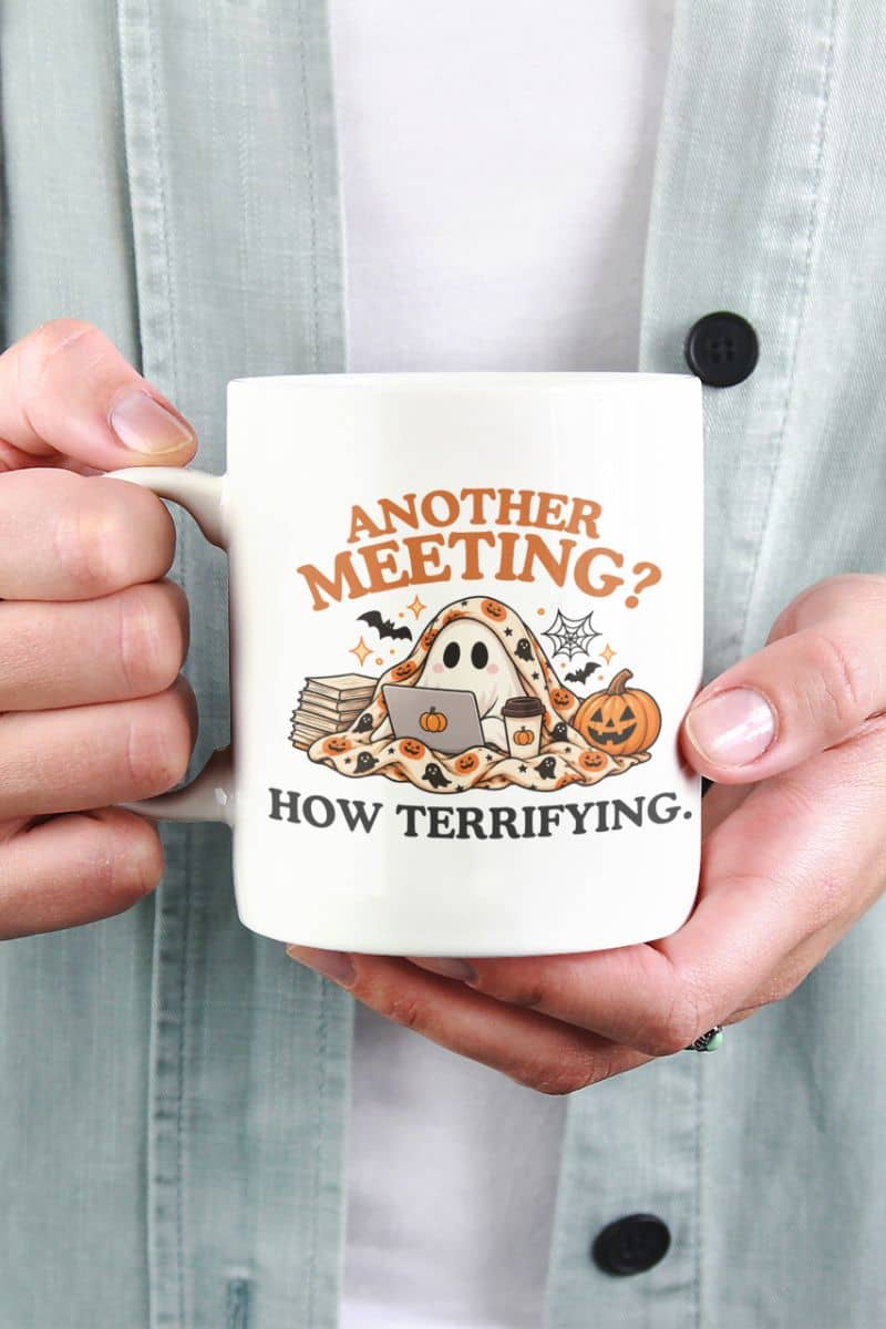 A person holding a meeting mug with a humorous Halloween graphic that reads 'Another Meeting? How Terrifying.', featuring a ghost, laptop, books