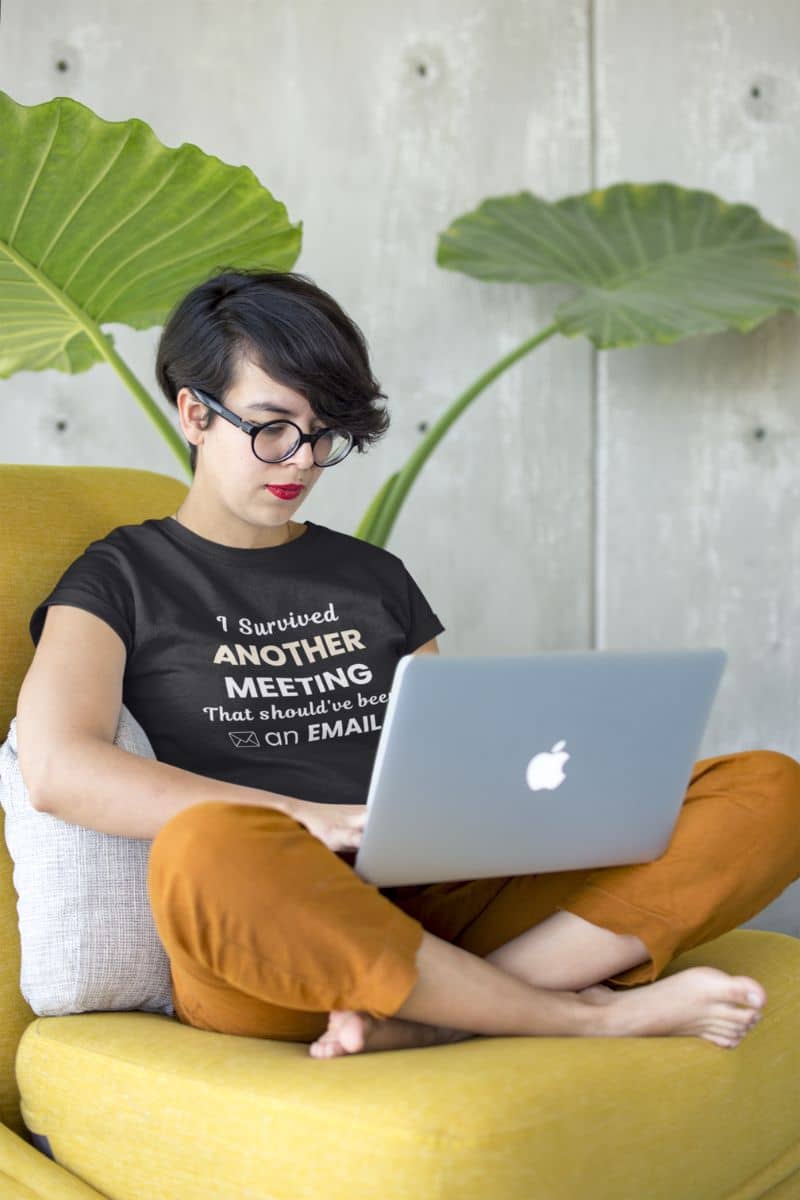 meeting survivor shirt with a woman wearing a black t-shirt featuring the text 'I Survived Another Meeting That Should’ve Been an Email', sitting on a yellow chair using a laptop with a plant in the background