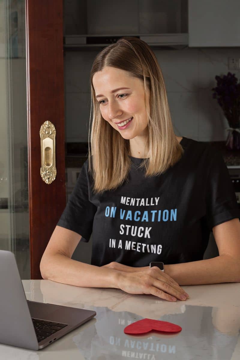 meeting survivor shirt featuring a woman wearing a black t-shirt with humorous text 'Mentally on Vacation. Stuck in A Meeting', sitting at a table with a laptop.