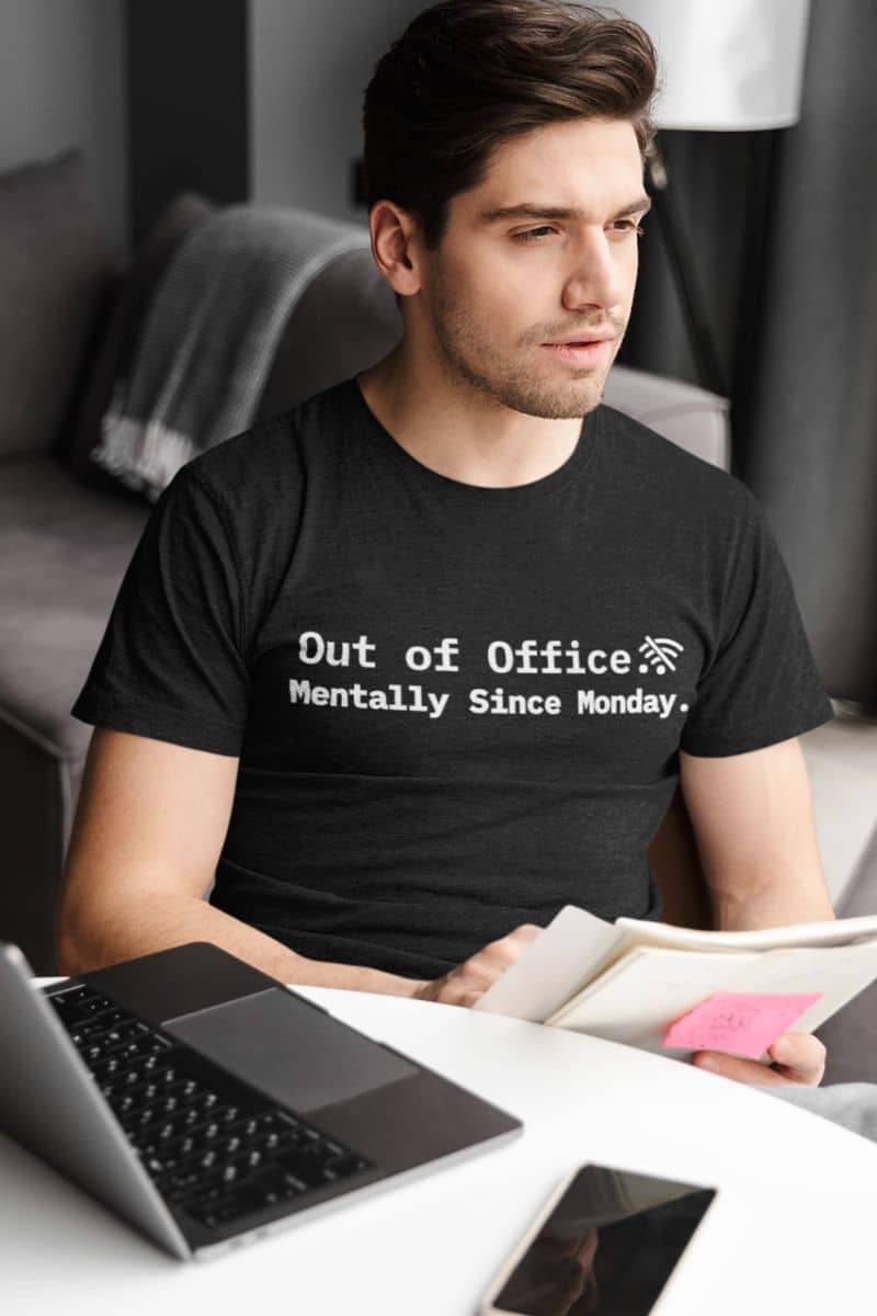 meeting survivor t-shirt with a man wearing a black t-shirt with text 'Out of Office. Mentally Since Monday.', sitting at a desk with a laptop and tablet.