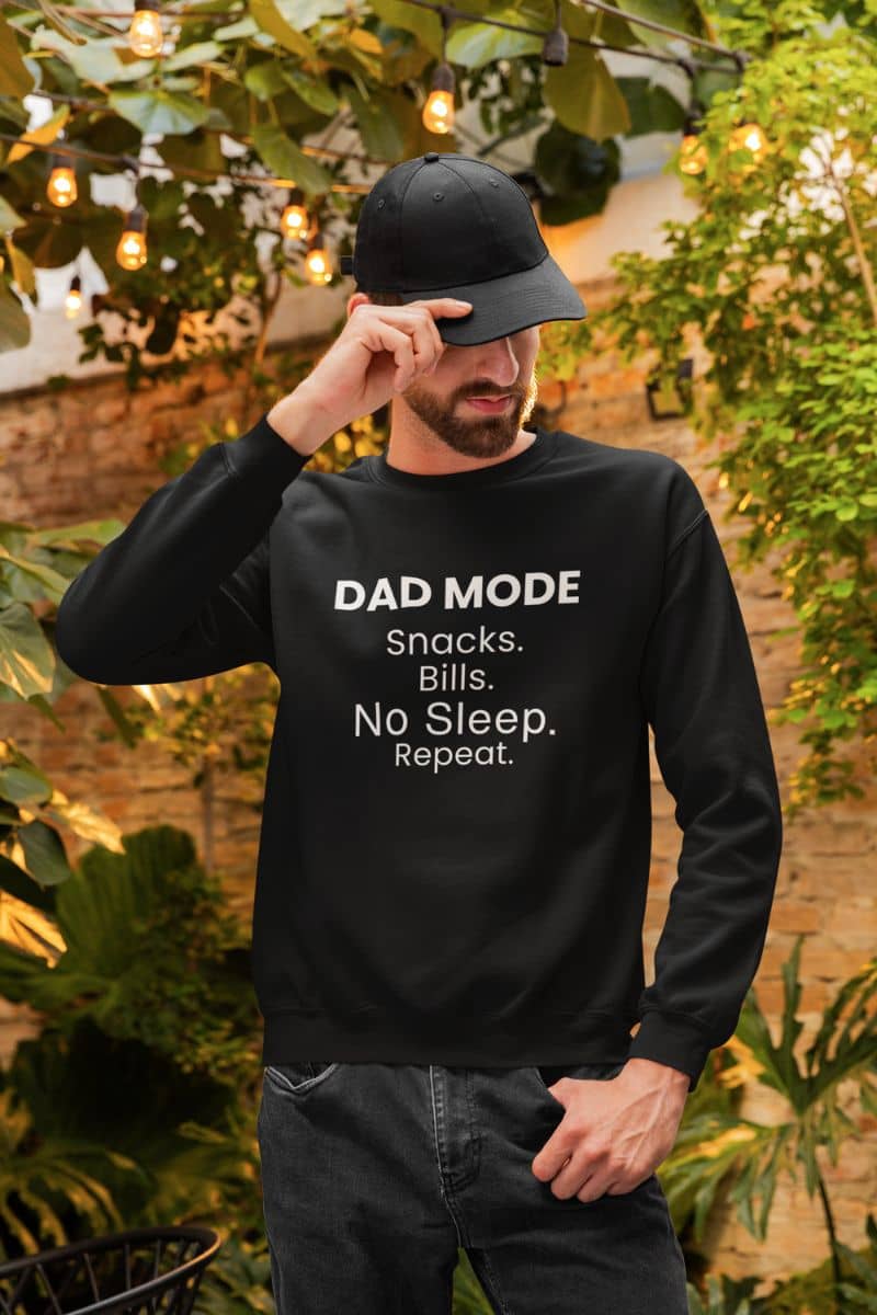 mens casual sweatshirt with a man wearing a black sweatshirt with 'DAD MODE: Snacks. Bills. No Sleep. Repeat.' text, standing outdoors with greenery.