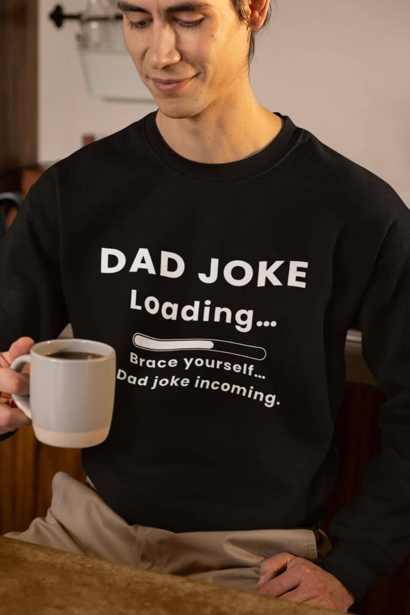 men's funny sweatshirt with a person wearing a black sweatshirt with 'Dad Joke Loading, Brace yourself…Dad joke incoming.' text, holding a mug.