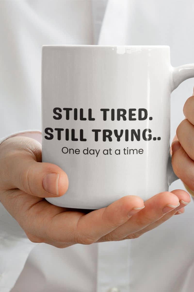 mental health reminder mug with a white mug with motivational text 'Still Tired. Still Trying. One day at a time', held by a person, against a light background
