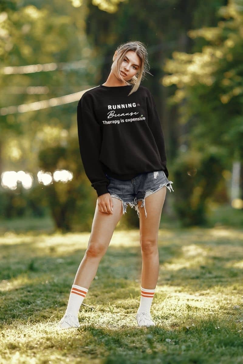 mental health running top with a person wearing a black sweatshirt with text 'Running because therapy is expensive', denim shorts, and white sneakers in a park.