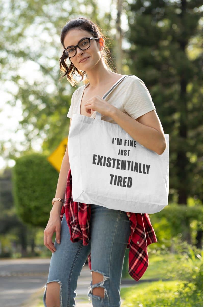 mental health tote bag with a woman holding a tote bag with text 'I’m fine. Just existentially tired.' outdoors 