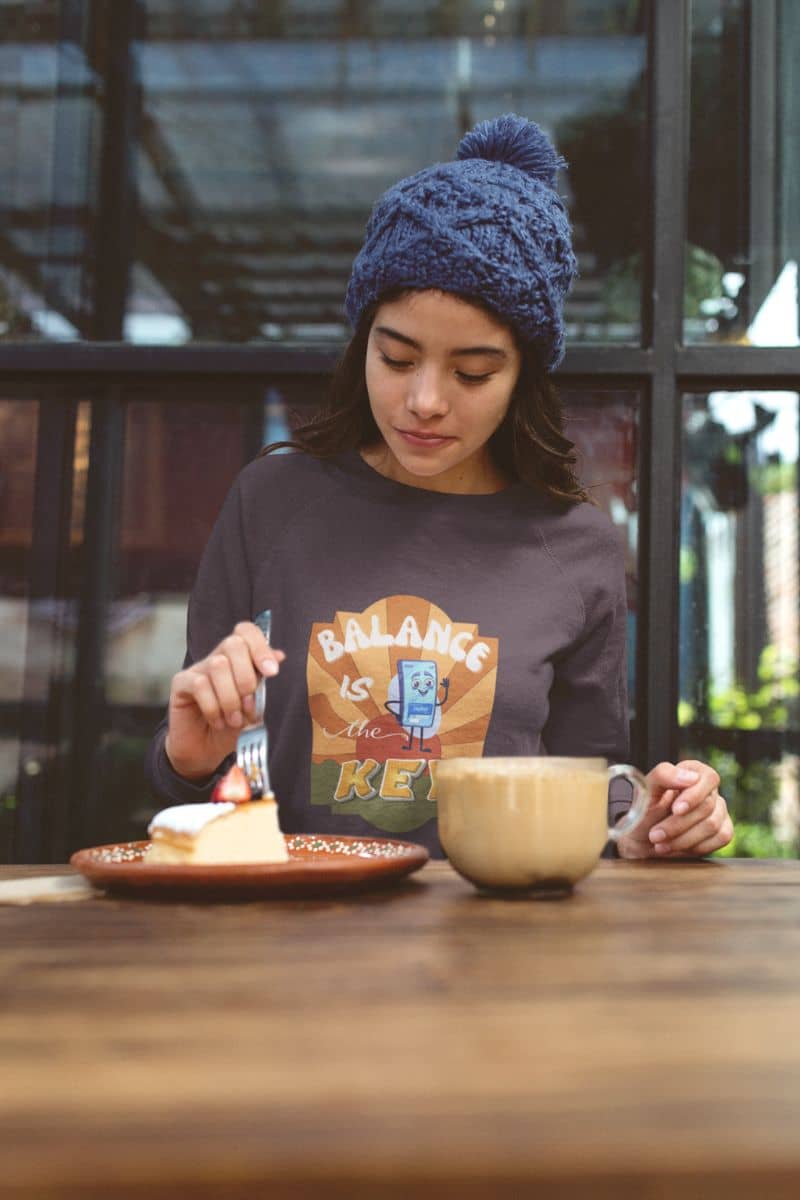 mental wellness pullover with a person wearing a blue knit hat and sweatshirt with a graphic design, sitting at a table with a plate of food and a mug.