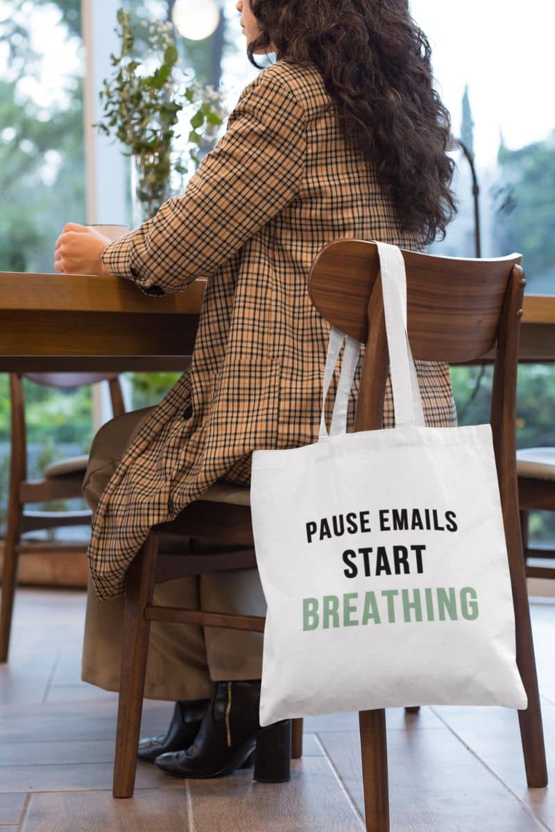 mental wellness tote with a person sitting at a table with a tote bag that says 'Pause Emails, Start Breathing' on a blurred background