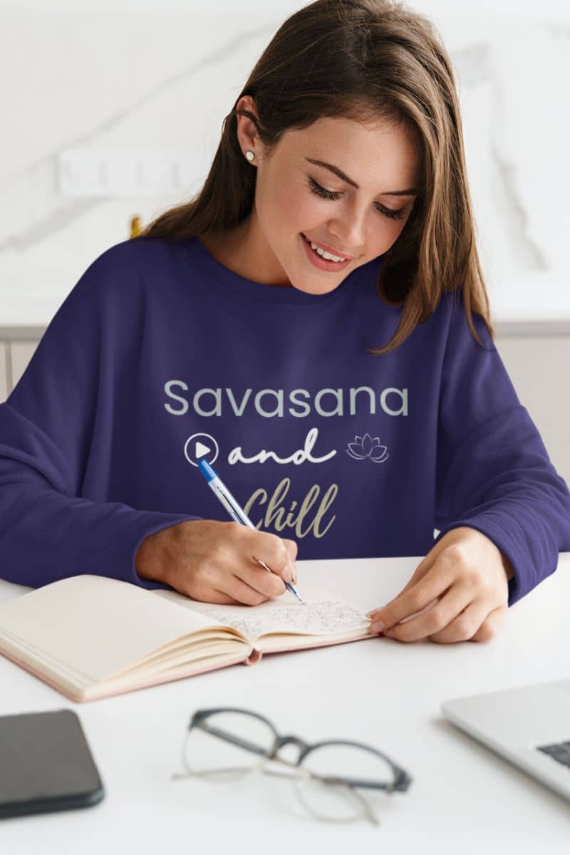 mindfulness sweater with a woman wearing a purple 'Savasana and Chill' sweatshirt, sitting at a desk with a notebook and pen.