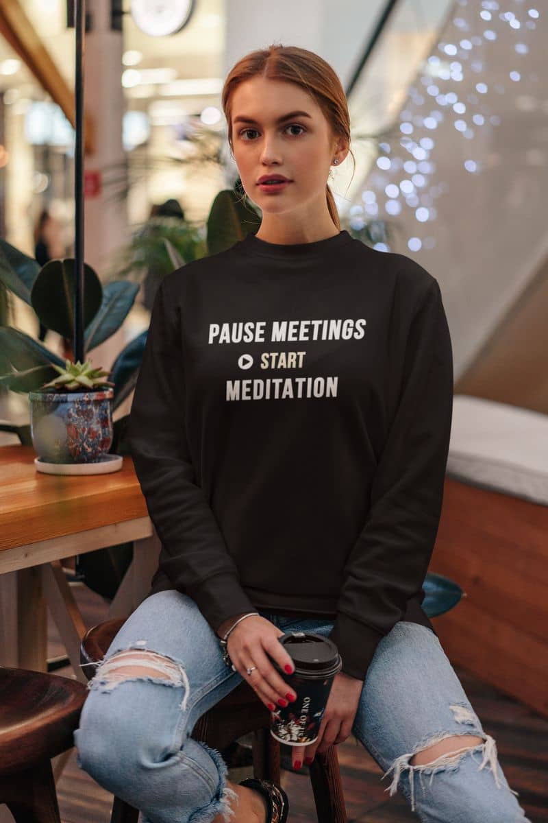 mindfulness sweatshirt with a woman wearing a sweatshirt featuring the text 'Pause Meetings, Start Meditation', while holding a cup of coffee in a coffee shop